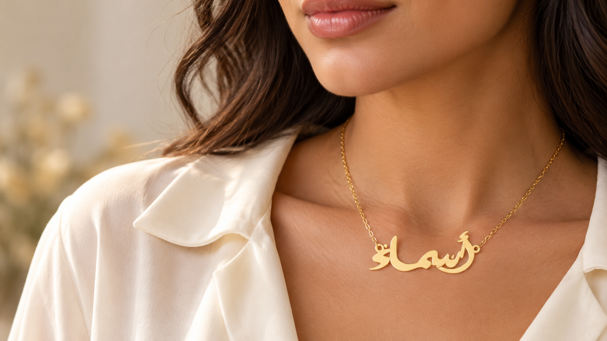 A close-up of a woman wearing a delicate gold necklace with an Arabic nameplate pendant, styled with a cream satin blouse.
